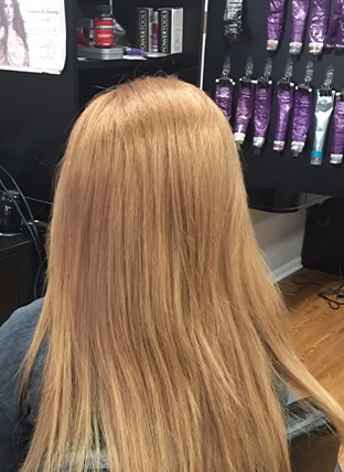 Back view of long straight blonde hair in a salon chair, with purple hair products on shelves in the background